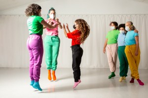 2 adults and one youth in a circle in the act of jumping up and clapping palms to the person next to them. 2 youths and one adult in the backround right corner with arms around each other and watching the jumpers. All in brightly colored sweats and T shirts.