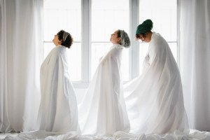 3 women in profile facing left wearing long white robes against a window in a light-filled white room. The first two on the left are gently arched back, looking up, the third is looking down.