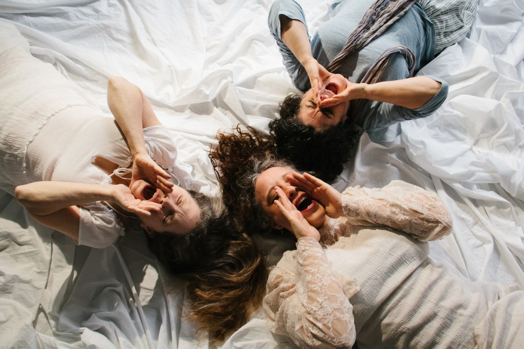 Shot from overhead: 3 women on backs, head to head, lying on white cloth. Their hands are on either side of their open mouths and they seem to be singing or shouting.